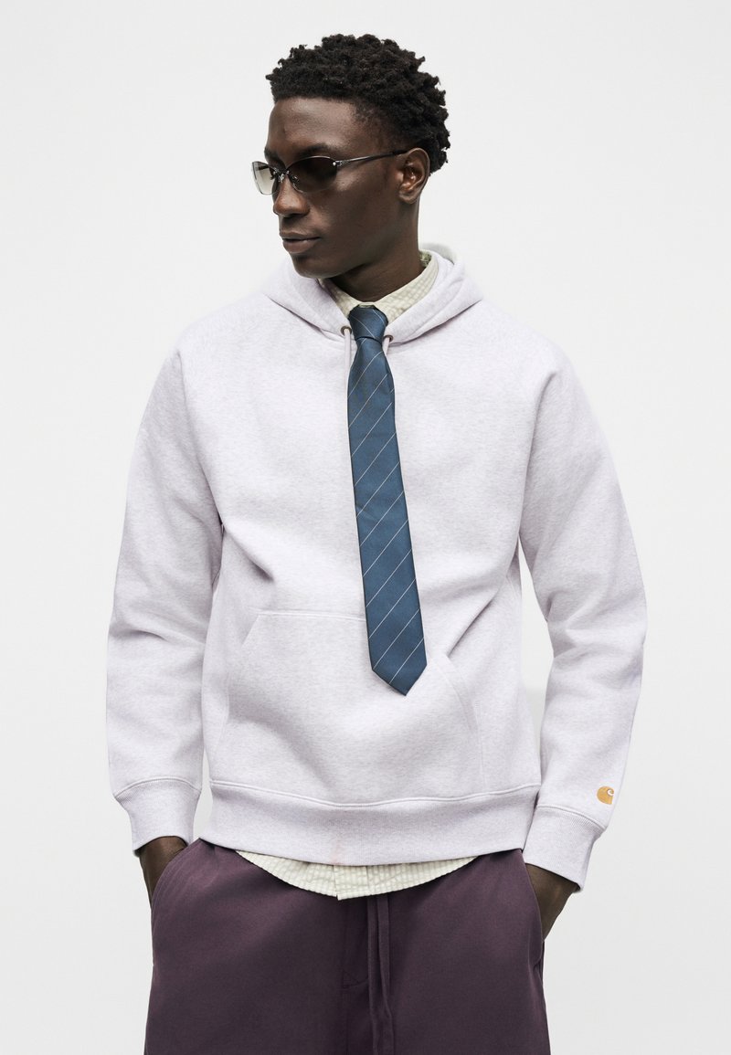 Jonge man met zonnebril, een lichtgrijze hoodie met een blauwe stropdas, geruit overhemd eronder en donkerpaarse broek, staand met handen in de zakken.