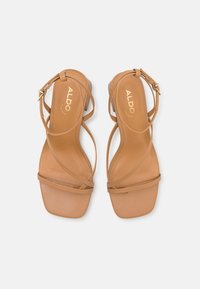 Beige høyhælte sandaler med tynne kryssede stropper og firkantet tå. Glatt lær-overdel med lav blokkhæl og gullhardware-accent.