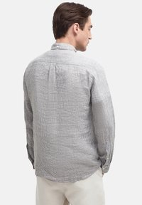 Mann in einem hellgrauen karierten Langarmhemd und beigen Hosen, der mit dem Rücken vor einem schlichten weißen Hintergrund steht.