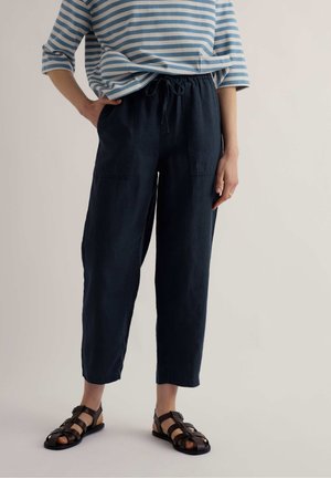 Osoba oblečená v námorníckych modrých nohaviciach so šnúrkou, modro-bielom pruhovanom tričku a čiernych sandáloch so remienkami stojaca pred jednoduchým pozadím.