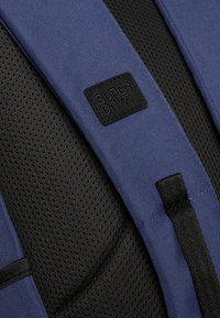 Tirantes de mochila de tela azul con acolchado de malla negra, que presentan un parche de logo en relieve de "SUPERDRY" en negro y acentos de costura negra duradera.