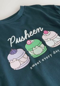 Mørk teal t-shirt med tre tegneserie-katte i makron-designs, i pastel-farver og teksten "Pusheen sweet every day" nedenfor.