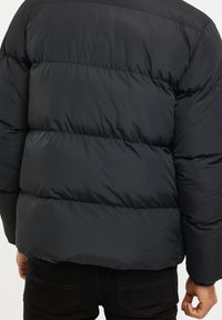 Veste matelassée noire portée par une personne vue de dos, avec un matelassage horizontal et un pantalon noir visible sous l'ourlet de la veste.