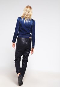 Donna bionda che si allontana indossando una giacca di jeans scura, pantaloni di pelle neri e sneakers nere su uno sfondo chiaro e semplice.