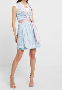 Femme portant une robe en dentelle bleu clair avec un ruban en satin rose et des sandales à talons hauts beiges à bout ouvert, debout devant un fond blanc.