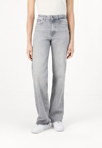 Personne portant un jean droit taille haute gris clair, des baskets blanches et un haut blanc rentré, debout devant un fond uni.