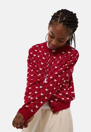 Cardigan rouge avec motif en losanges, featuring de petits coeurs blancs. Manches et ourlet côtelés, fermeture par boutons et texture douce. Porté par-dessus un top blanc.
