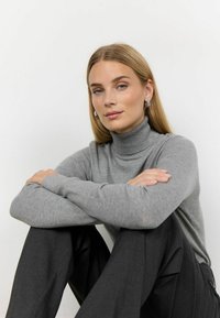 Šedý rolák vyrobený z měkkého materiálu, přiléhavé dlouhé rukávy, nošený s vysokým pasem černými kalhotami, v sedící poloze se zkříženýma rukama.