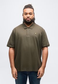 Άνδρας με πλεξούδες dreadlocks που φοράει μια ελιά πράσινη πόλο μπλούζα και μπλε τζιν, στέκεται μπροστά σε ένα απλό λευκό φόντο, αντιμετωπίζοντας προς τα εμπρός.