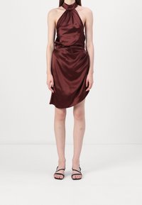 Femme portant une robe en satin bordeaux sans manches à col licou et des sandales noires à brides, debout devant un fond blanc uni.