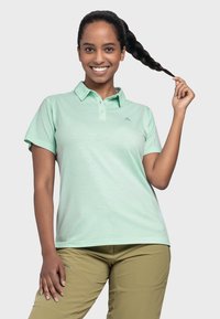 Hellgrünes Poloshirt mit Kragen, kurzen Ärmeln und einem dezenten Logo auf der Brust. Glatte Textur, kombiniert mit hellbraunen Hosen.