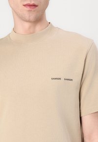 Beige, rövid ujjú póló magas nyakkal. Két fekete, hímzett "SAMSØE" logó található a mellrészen. Puha pamut anyagból készült.