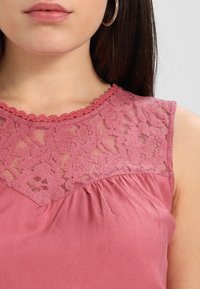 Top senza maniche di colore rosa con yoke in pizzo e motivi floreali, dettagli arricciati sotto il colletto e una consistenza morbida.