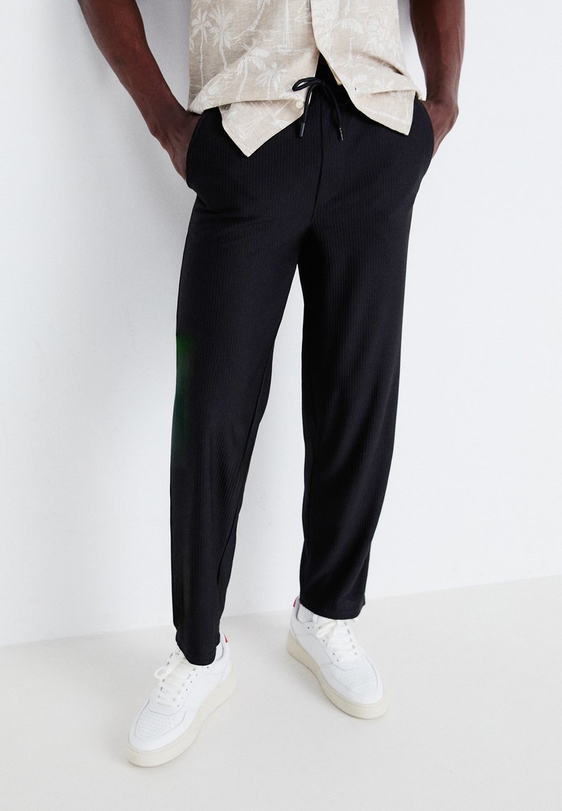 Black, ribbed jogger pants with an elastic waistband and drawstring, paired with white sneakers. The fabric appears lightweight and casual.