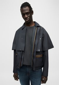 Αδιάβροχο, μπουφάν με κουκούλα σε σκουρό γκρι χρώμα με κουμπιά τύπου κουμπί και πορτοκαλί λεπτομέρεια. Διαθέτει υπερμεγέθη σχεδιασμό και πολλαπλές τσέπες.