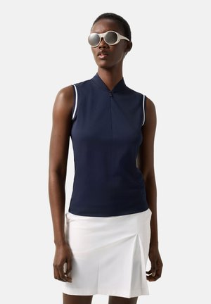 Woman wearing white pleated skirt and navy sleeveless top with zipper collar, accessorized with large white sunglasses.