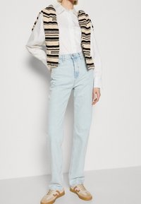 Ljusblå högmidjade jeans, parat med en vit skjorta med knappar och en svartstripad, krämfärgad stickad väst. Bruna och tan sneakers.