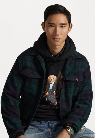 Tamsiai žaliai ir mėlynai dryžuota flanelinė striukė, uždėta ant juodos džemperio su lokio grafika. Matomi nusidėvėję džinsai ir laikrodis.