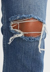 Los pantalones de mezclilla azul presentan un gran desgarro desgastado en la rodilla con bordes deshilachados, exponiendo la piel debajo. La textura es suave y usada.