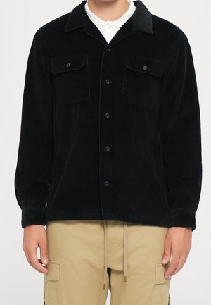Mann trägt schwarze Cordjacke mit Knöpfen und zwei Brusttaschen sowie beige Cargohose mit Kordelzug am Bund.