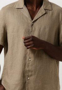 Persona di pelle scura che indossa una camicia di lino beige a maniche corte con bottoni, con la mano alzata vicino al petto su uno sfondo neutro.