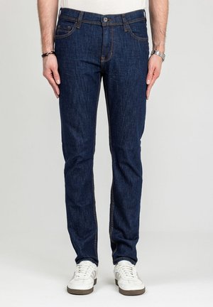 Mężczyzna ubrany w ciemnoniebieskie dopasowane jeansy i białe trampki stojący na jasnym, jednolitym tle.