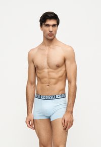 Modèle masculin aux cheveux foncés portant un boxer bleu clair de la marque Diesel, debout devant un fond blanc uni.