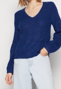 Głęboki niebieski sweter w serek wykonany z miękkiej, strukturalnej dzianiny z długimi rękawami. Połączony z jasnoniebieskimi dżinsami, podkreślającymi swobodny styl.