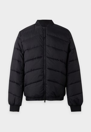 Jeune homme portant une veste matelassée noire sur un pull en maille blanc, un jean bleu et des baskets rouges, debout avec une main dans la poche.