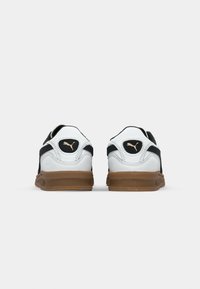 Paire de baskets Puma blanches et noires avec semelles marron vues de dos sur un fond uni gris clair.