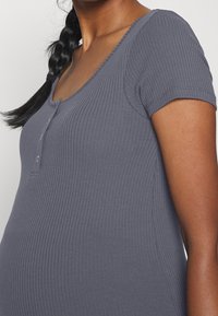 Persona embarazada que lleva una camiseta ajustada de manga corta y con canalé de color gris, con botones cerca del escote y un peinado de trenza lateral.