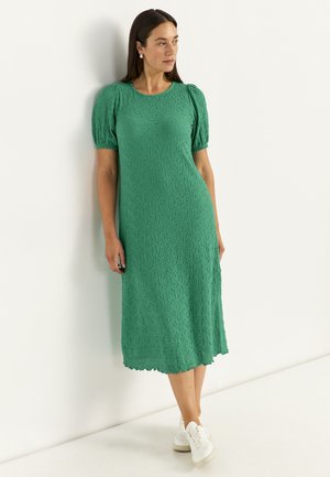 Kvinde iført en tekstureret grøn midi-kjole med puffede ærmer og hvide sneakers, stående op ad en ensfarvet hvid væg.