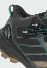 Czarno-zielone buty trekkingowe z teksturowaną siateczką, wyraźnym logo marki, grubą gumową podeszwą i odblaskowymi akcentami oraz zapięciem na sznurówki.