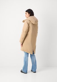 Lys brun, lang polstret jakke med hette, med en splitt bak og justerbar snor, båret over blå vide jeans.