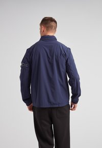 Chaqueta ligera azul marino con cuello alto, puños elásticos y un bolsillo con cremallera en la manga; tejido suave y resistente al agua.