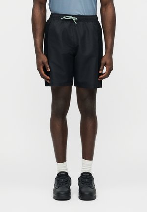 Férfi fekete sportnadrágot visel oldalán hálós panelek, világoskék póló, fehér zokni és fekete sportcipő, fehér háttér előtt áll.