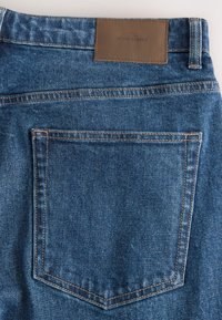 Gros plan sur la poche arrière d'un jean en denim bleu avec des coutures visibles et une étiquette en cuir marron portant la mention "DENIM SUPPLY" sur la ceinture.