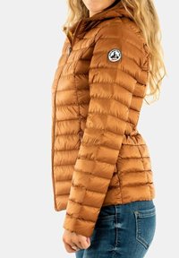 Doudoune marron avec texture matelassée, dotée d'une capuche, d'une fermeture éclair et d'un patch logo sur l'épaule gauche. Portée avec un jean bleu.