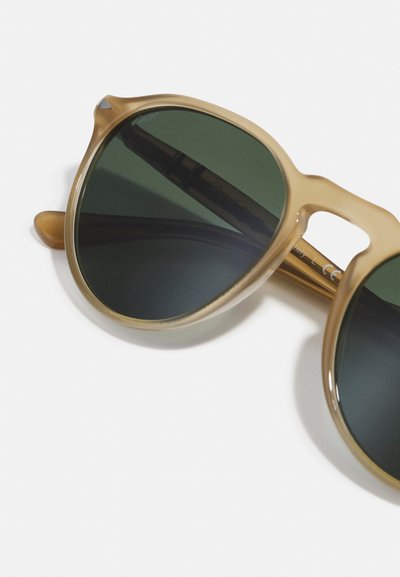 Ochelari de soare cu un cadru transparent de nuanță bej, lentile de un verde închis, formă rotundă și un accent argintiu în zona tâmplei. Textura netedă.