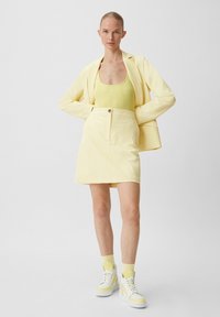 Gelber Blazer über einem passenden gelben Tanktop, kombiniert mit einem gelben Rock. Weiße Sneakers mit gelben Akzenten und weißen Socken runden den Look ab.