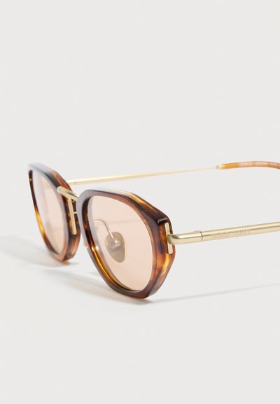 Γυαλιά ηλίου τύπου γατίσια μάτια με σχέδιο κέλυφος χελώνας, χρυσά μεταλλικά μπράτσα και φακούς με ανοιχτό καφέ απόχρωση σε λευκό φόντο.