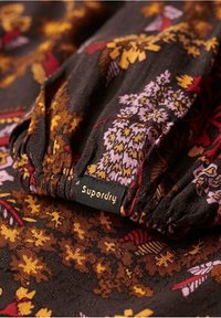 Tissu à motifs floraux dans des tons foncés avec des accents jaunes, violets et rouges ; présente un col froncé et une étiquette Superdry visible.