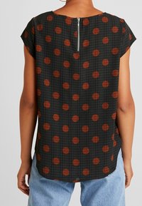 Haut noir à manches courtes avec des pois rouges et un motif texturé, dotée d'une fermeture éclair au dos et d'un ourlet arrondi.