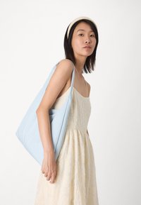 Bolsa tote de color azul claro con una textura suave, diseño minimalista y asas anchas, sostenida contra un vestido color crema con patrones florales.