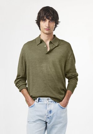 Młody mężczyzna o ciemnych, falowanych włosach, ubrany w oliwkowo zieloną koszulkę polo z długim rękawem i jasnoniebieskie dżinsy, stoi z rękami w kieszeniach.