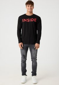 Camisa negra de manga larga con letras rojas que dicen "INSIDE", combinada con jeans grises desgastados y zapatillas blancas, presentando una silueta ajustada.