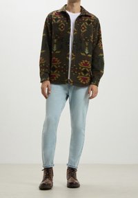 Koszula flanelowa w wielokolorowy wzór z kołnierzykiem, z dwiema przednimi kieszeniami, noszona na białym t-shircie, w zestawie z jasnoniebieskimi dżinsami i brązowymi butami.
