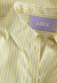 Camicia a righe gialle con colletto, caratterizzata da righe bianche e grigio chiaro, chiusura con bottoni e un'etichetta viola con la scritta "JJXX."
