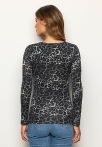 Frau mit welligem braunem Haar, die ein langärmeliges, schwarz-graues Leopardenmuster-Oberteil und eine blaue Jeans trägt und mit dem Rücken zu einem schlichten Hintergrund steht.