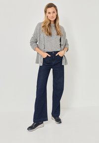 Siva pletena jopica preko sive majice, v kombinaciji z temno modrimi širokimi hlačami in črnimi teniskami z belim podplatom. Ohlapen outfit.
